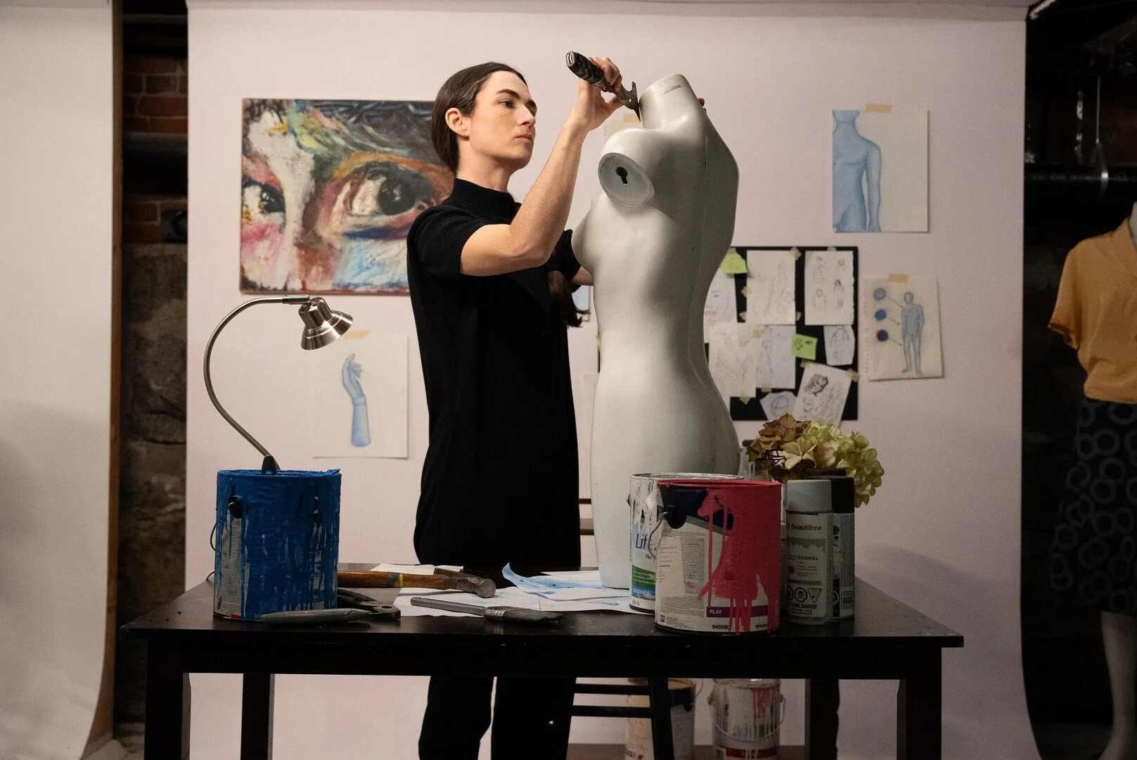 Woman painting a mannequin torso in an art studio with paintings and sketches displayed on the wall behind her.