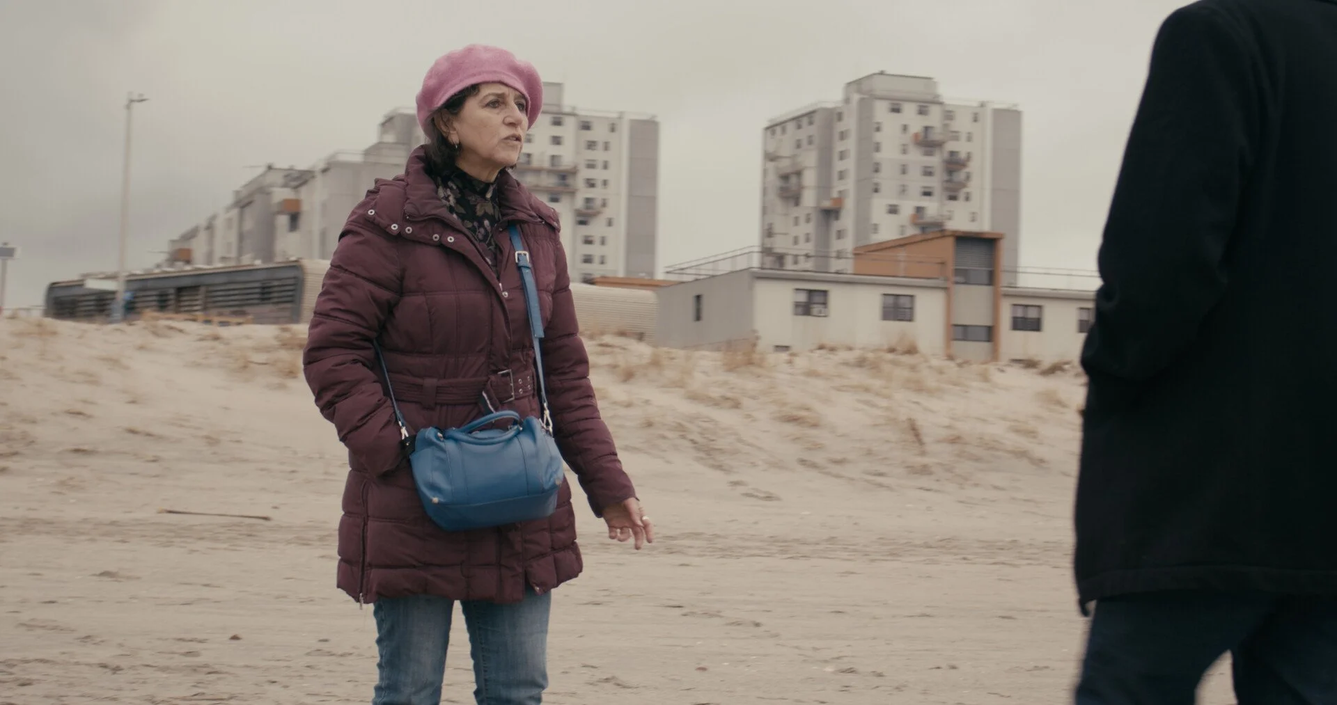 Older woman standing on sandy beach, wearing a pink hat and a maroon jacket, speaking to a man dressed in black with blurred city buildings in the background.