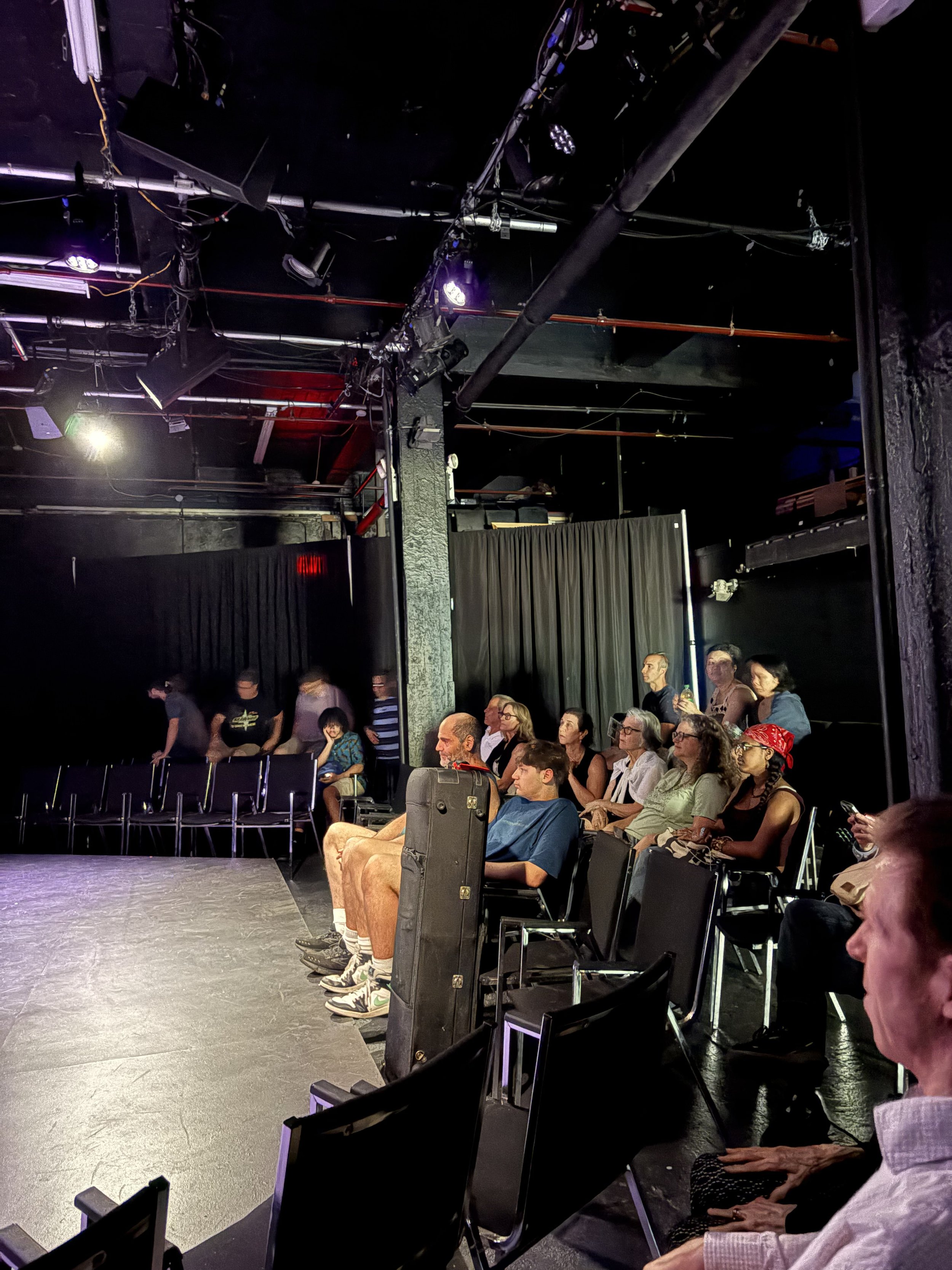 Audience sitting in black chairs in a small theater or comedy club, watching a performance on stage. The stage is empty and black curtains are visible in the background. The room is dimly lit with some stage lighting overhead.