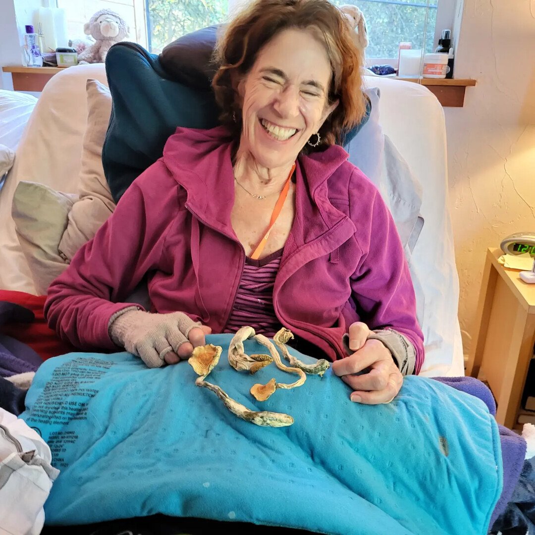 A cheerful woman with short brown hair, wearing a purple zip-up jacket and striped shirt, sitting in a hospital bed with dried mushrooms on her lap, smiling widely.