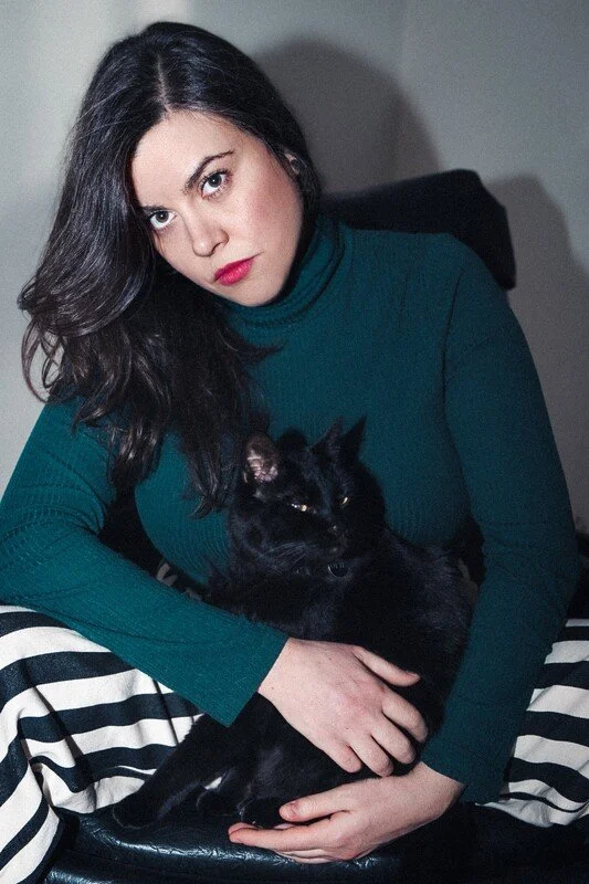 A woman with dark hair and red lipstick sitting on a black and white striped chair, holding a black cat in her lap.
