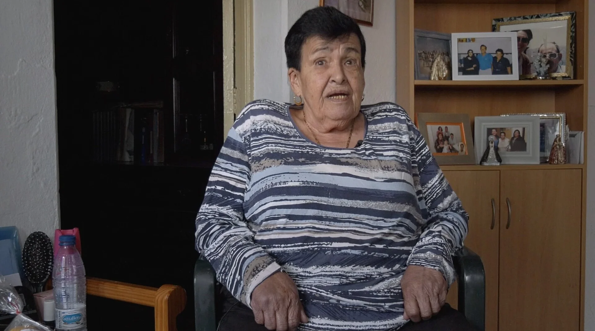 An elderly woman sitting in a black chair in a cozy room, wearing a patterned long-sleeved shirt, with a wooden bookcase with framed photographs behind her.