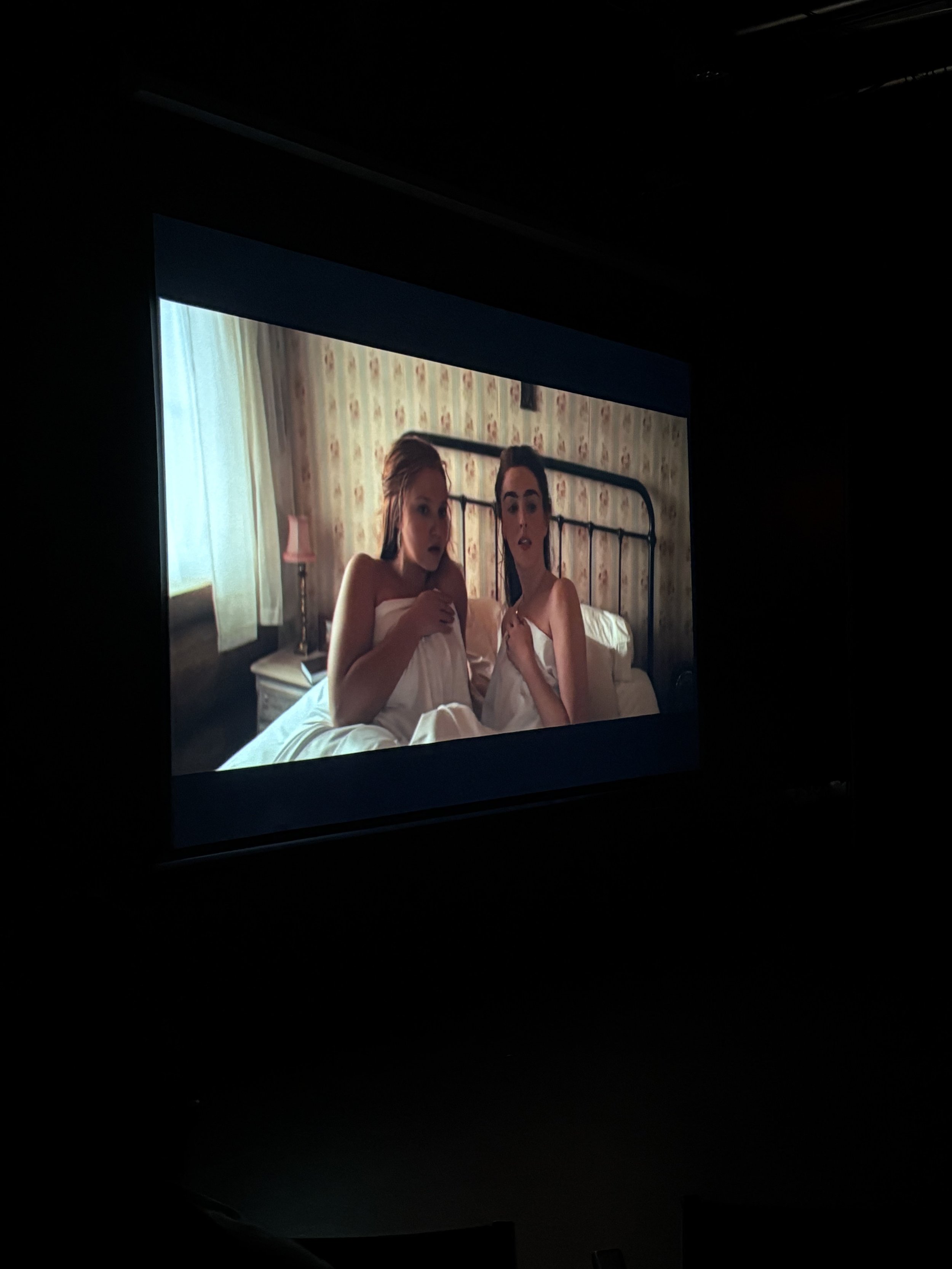 Two women sitting up in bed, wrapped in white sheets, looking surprised while watching a television screen in a bedroom with floral wallpaper, a window with curtains, and a bedside lamp.