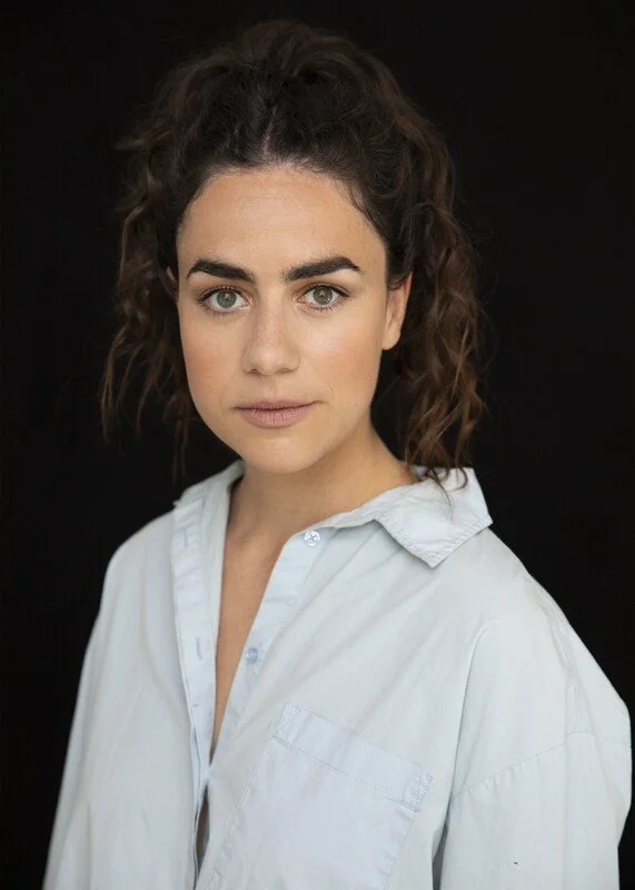 Portrait of a woman with dark curly hair, light skin, and green eyes, wearing a white button-up shirt against a black background.