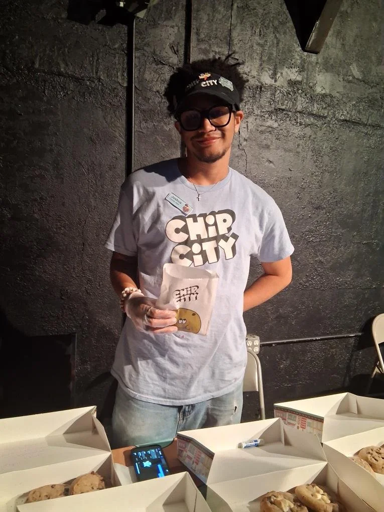 A young man with curly hair, wearing large sunglasses, a black cap, and a gray T-shirt with 'Chip City' printed on it, stands behind a table with boxes of cookies at a bake shop or bakery. He is holding a bag of cookies and smiling.