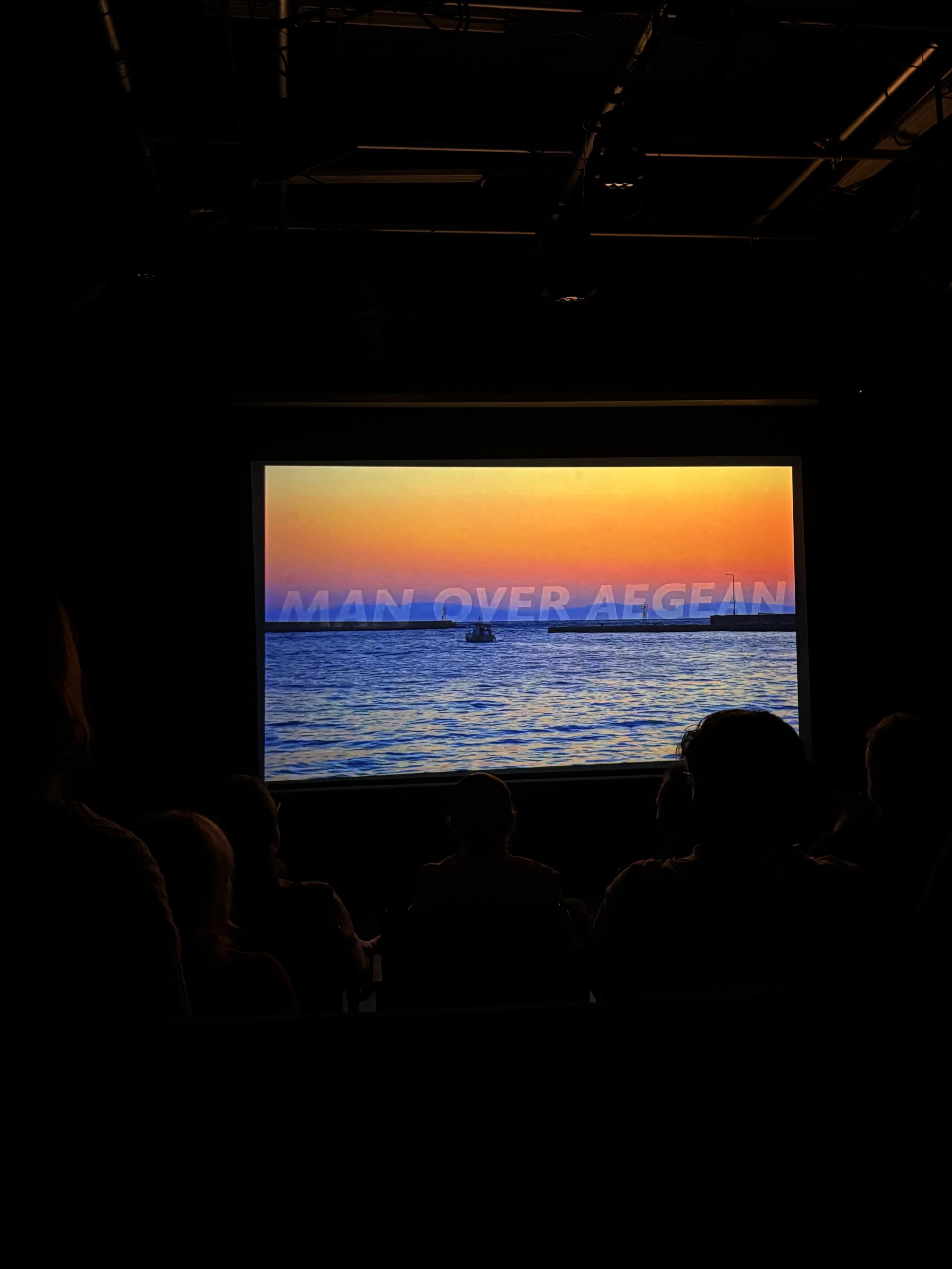 Audience watching a presentation with a large screen displaying a sunset over water and the words 'MAN OVER AEGEAN'