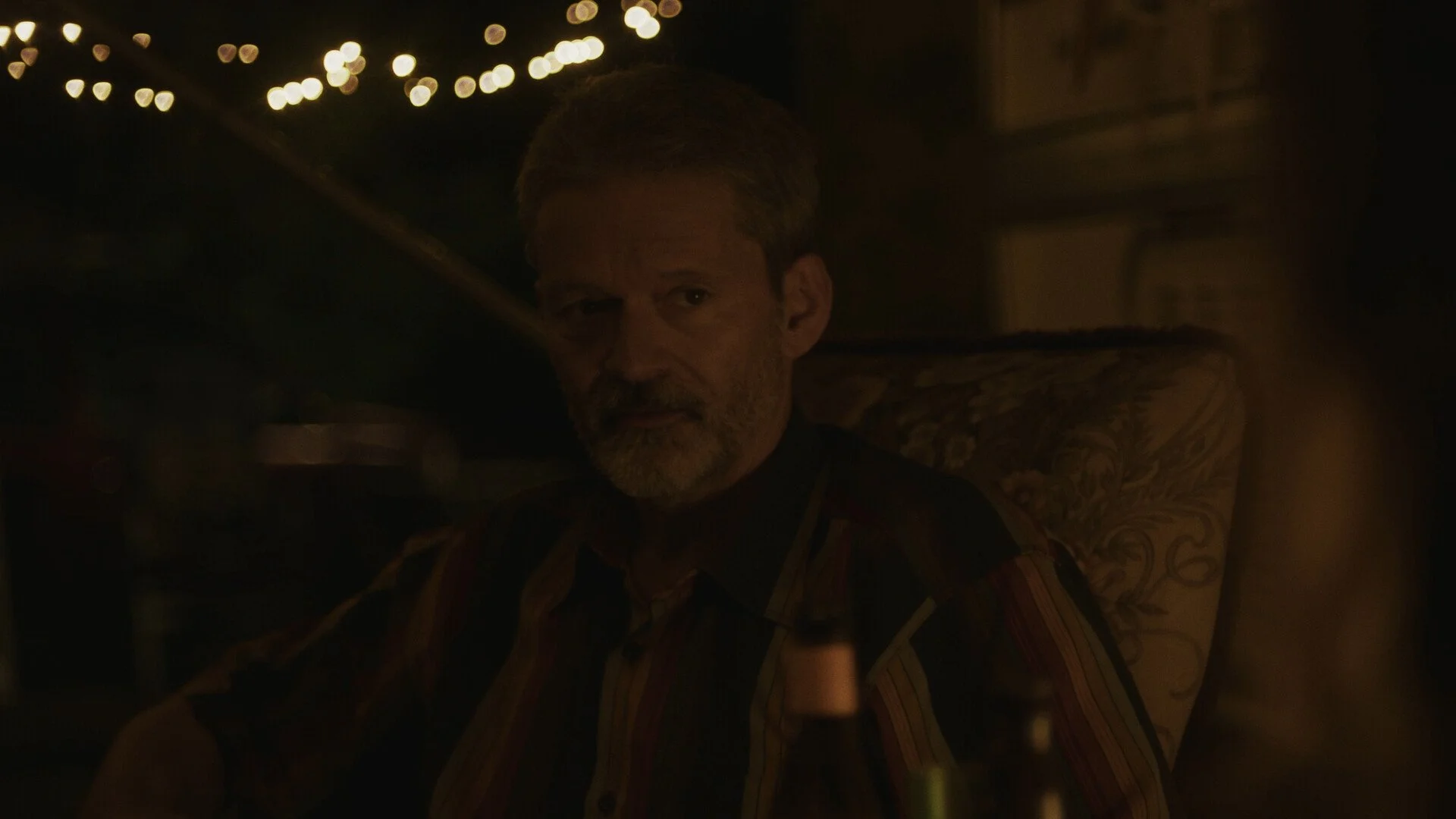 A man with gray hair and a beard sitting on a patterned armchair in a dimly lit room, with string lights in the background.