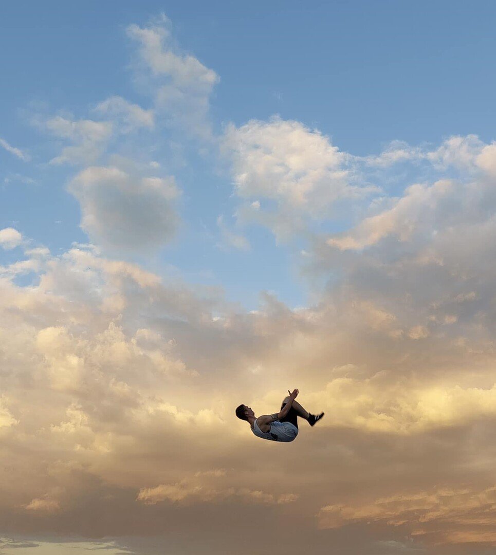 A person appears to be floating or falling in mid-air against a backdrop of a colorful sunset sky with clouds.