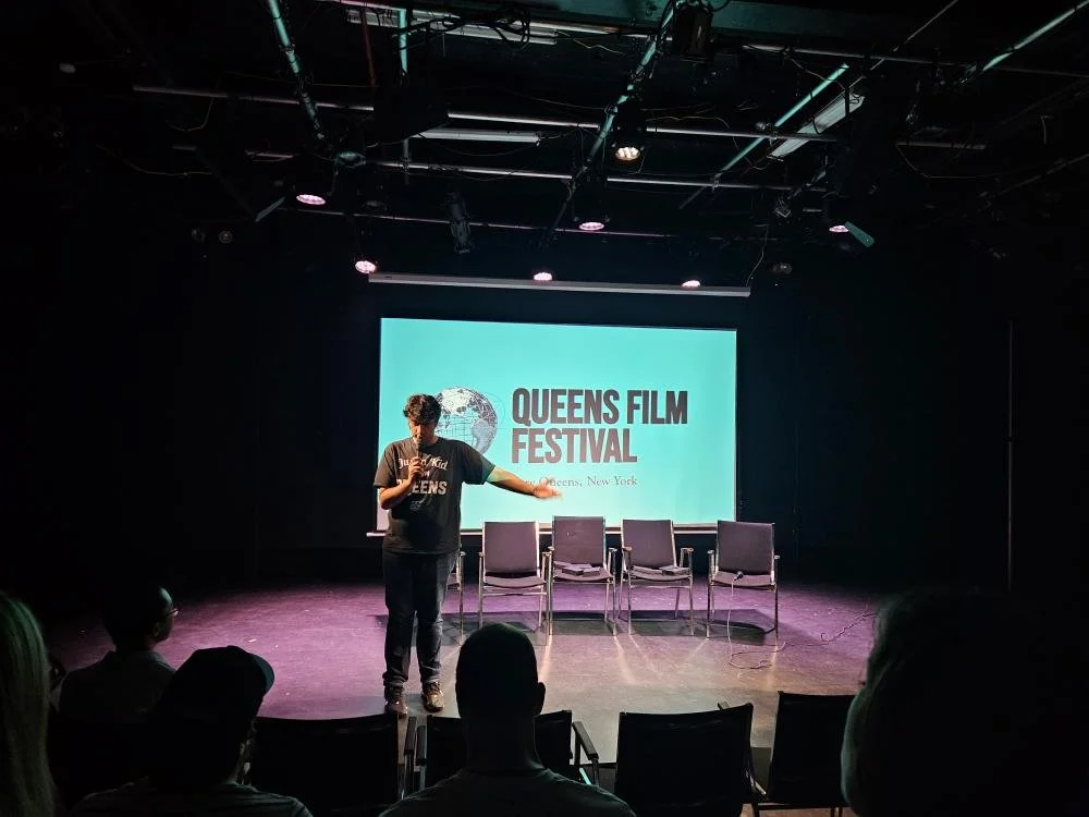 A person speaking on stage at the Queens Film Festival, with a presentation screen displaying the event's logo and name behind them. Audience members are seated facing the stage.