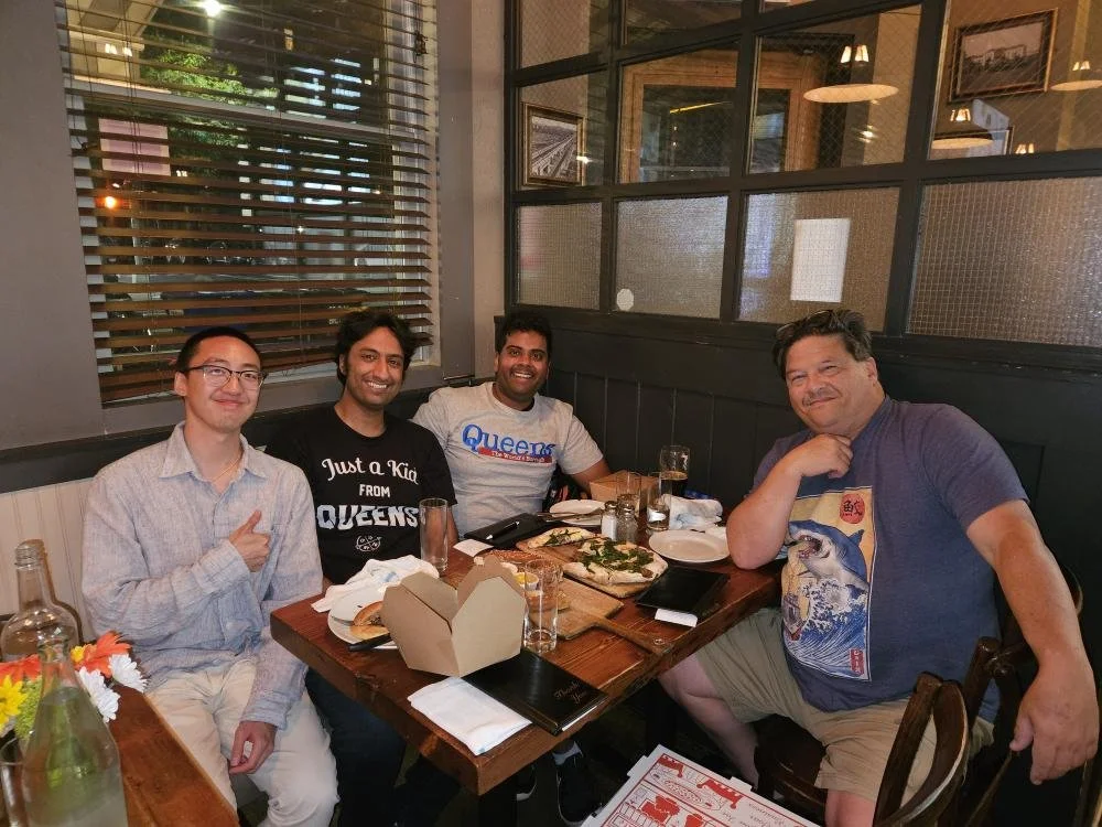 Four men sitting at a restaurant table with pizza and drinks, smiling and enjoying their meal.