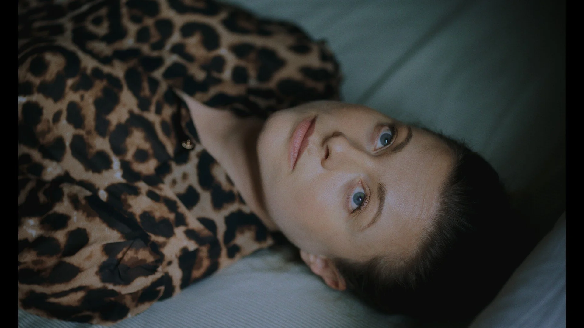 A woman with blue eyes and dark hair lying on a pillow, wearing a leopard print top.