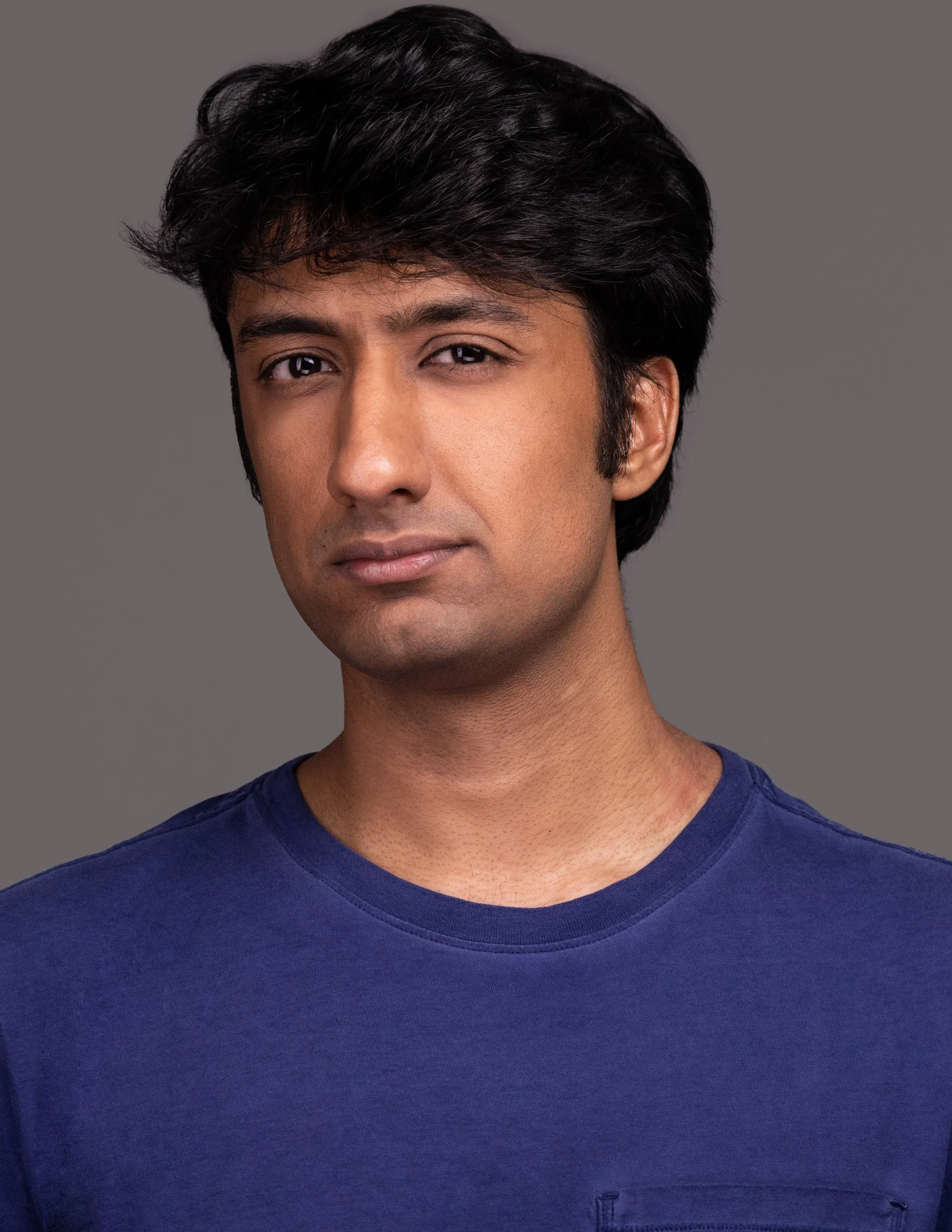 A man with black hair and medium skin tone wearing a blue t-shirt, looking directly at the camera with a neutral expression against a gray background.