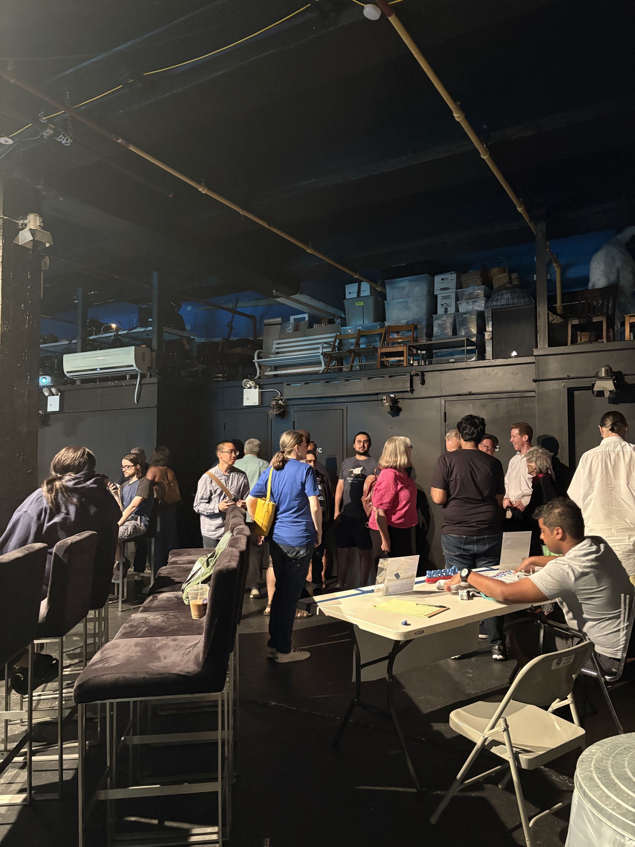 People gathered in a dimly lit indoor space, with some seated at a table and others standing in small groups, possibly at a social or networking event.