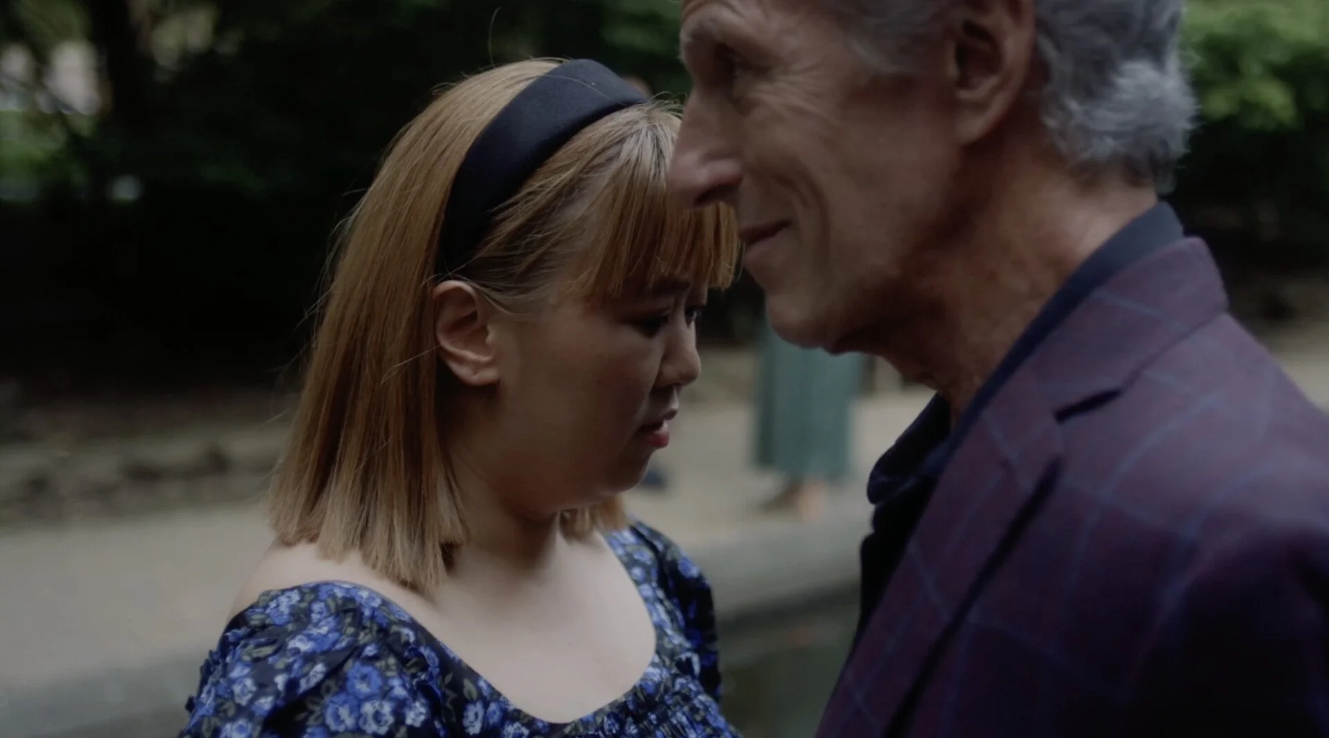 A woman and an older man are standing close together outdoors, with their foreheads touching and eyes closed, sharing an intimate moment.