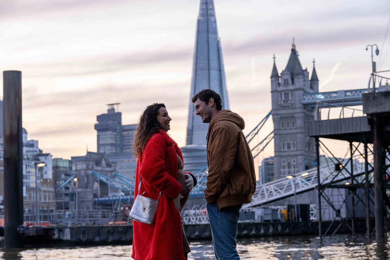 A smiling couple standing by the river with the London Bridge and the Shard in the background during sunset.