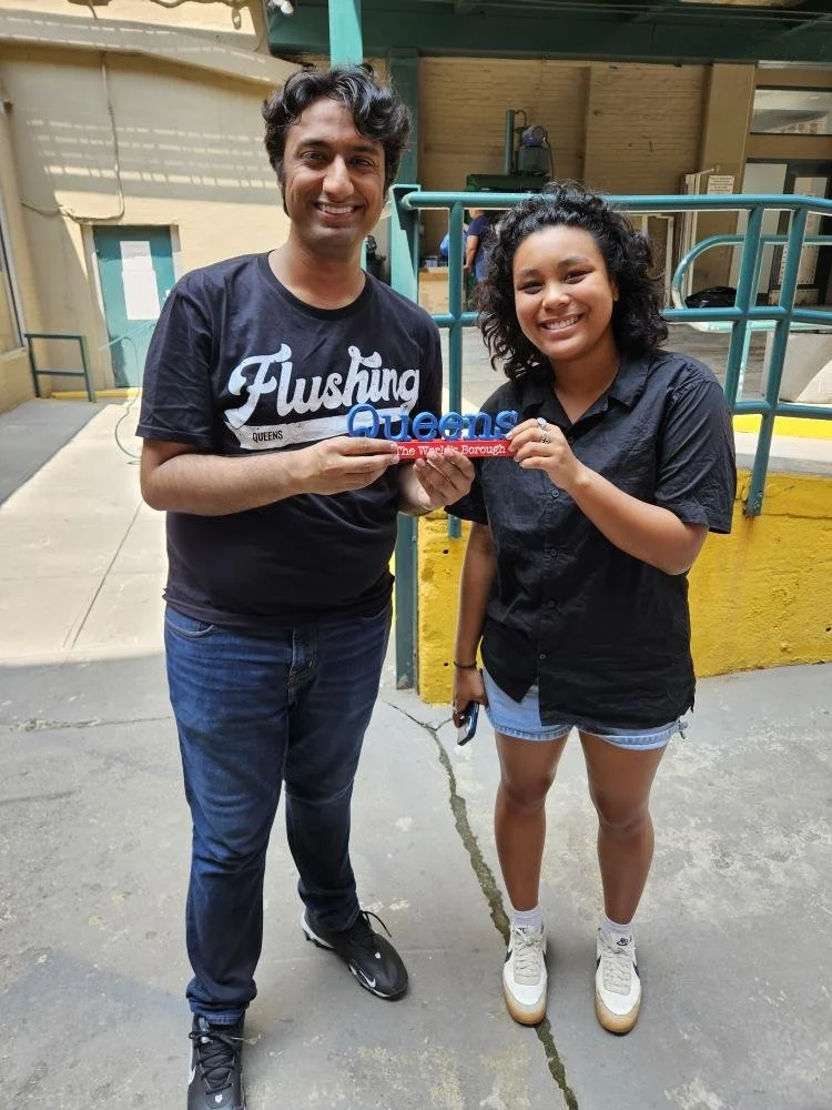 A man and a woman are smiling and holding a sign that reads "Queens" with extra text underneath. They are standing outside on a concrete surface near a yellow and teal railing.