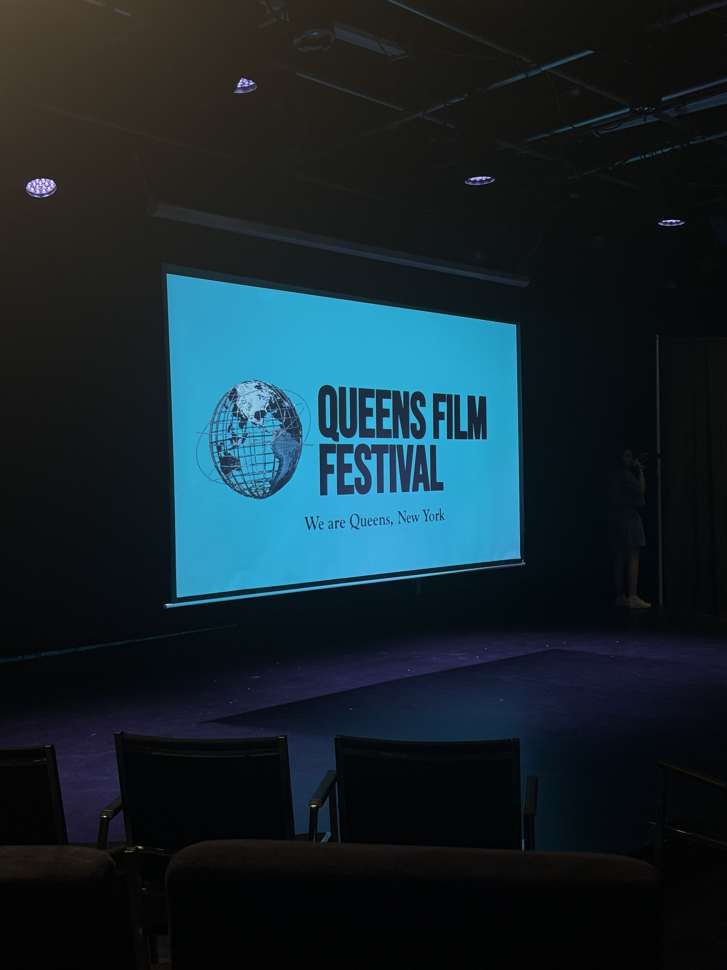 An indoor stage with a large screen displaying the Queens Film Festival logo with a globe and the text "Queens Film Festival" and "We are Queens, New York." There are chairs in front of the stage and a person standing to the right.