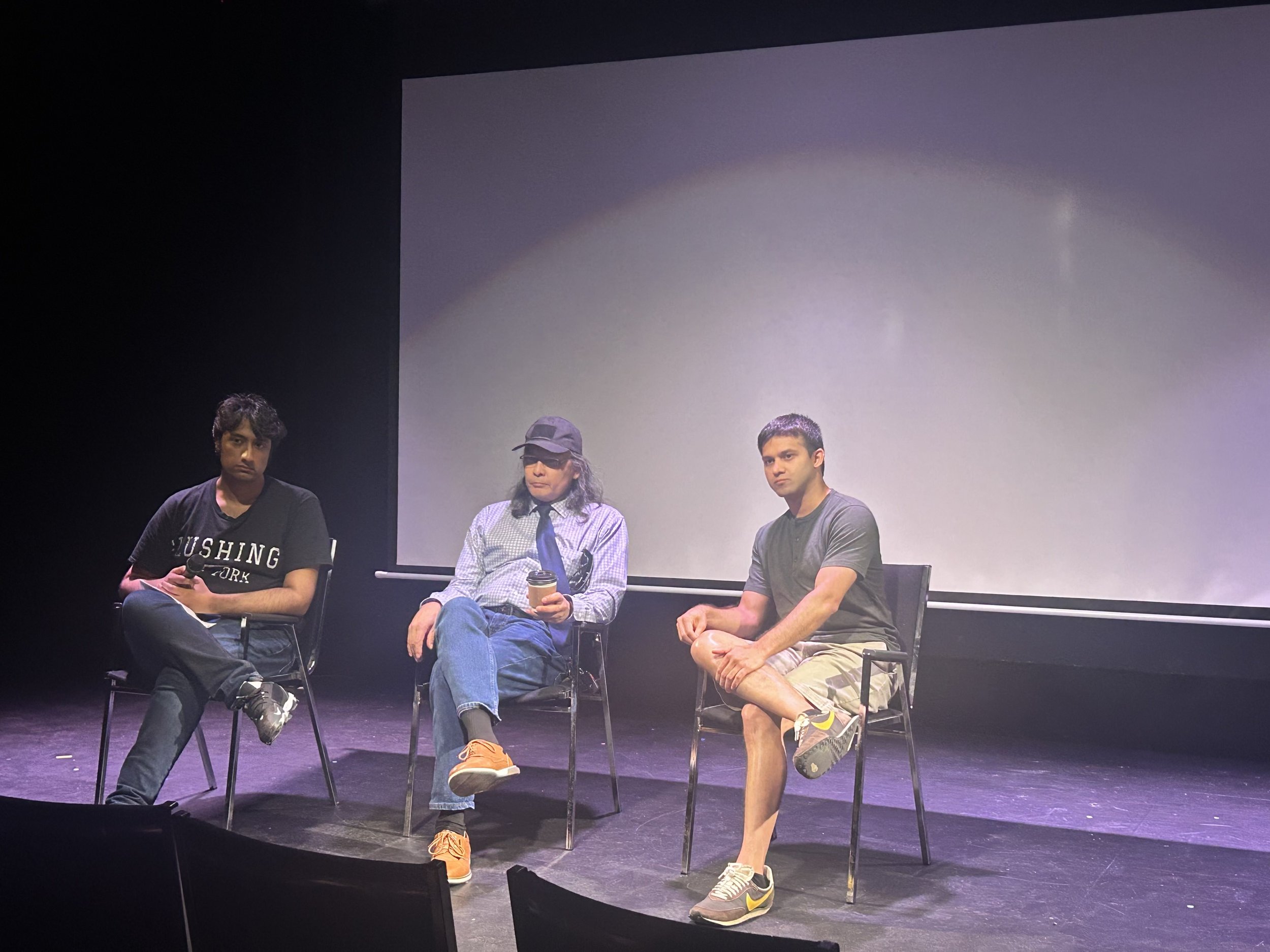 Three men sitting on chairs on a stage with a blank screen behind them, during a panel or discussion event.