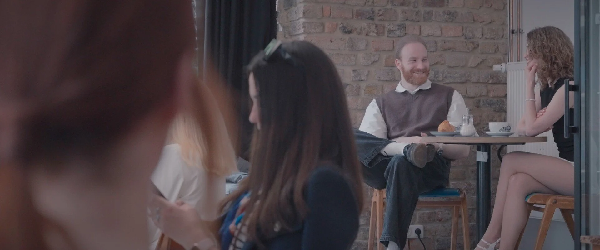 People sitting at a small table in a cozy cafe, smiling and engaged in conversation. The background features a brick wall.