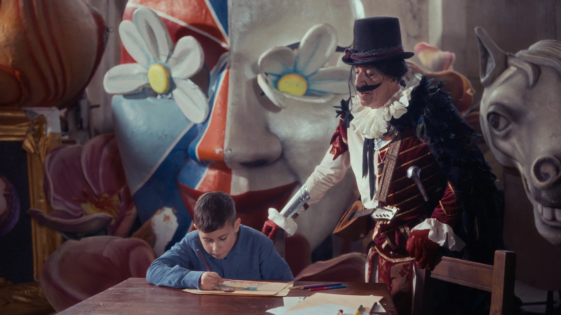 A young boy sitting at a table drawing or coloring, with a figure dressed as a clown or carnival character standing next to him in front of large colorful sculptures of flowers, faces, and a horse head in the background.