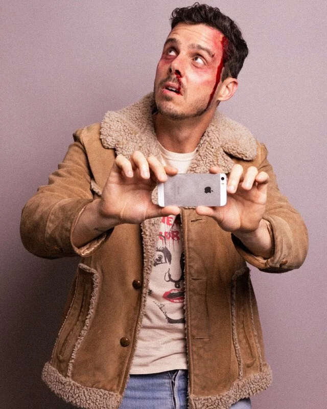 A man with a head injury, blood streaming down the side of his face, is holding a smartphone and taking a selfie while looking up, wearing a tan jacket with a furry collar.