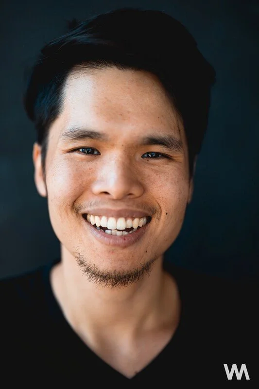 A young man with dark hair, light skin, and a beard, smiling against a black background.