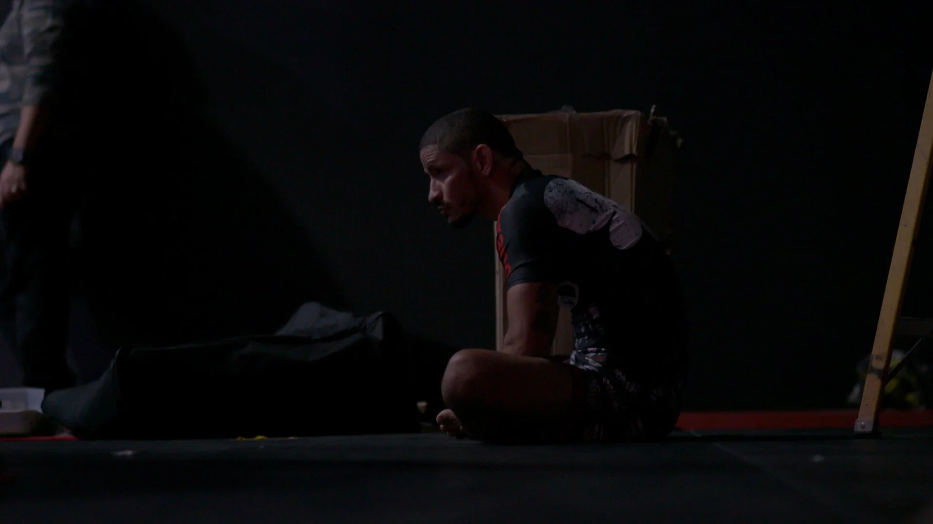 A man sitting on the floor in a dark room, wearing athletic clothing, with a cardboard box behind him.