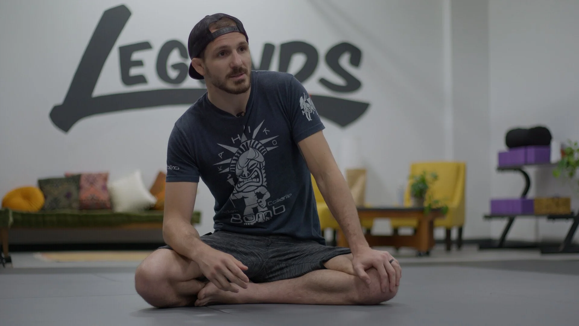 A man sitting cross-legged on a yoga mat, wearing a navy t-shirt and a backward baseball cap, in a room with a large wall sign reading 'LEGENDS' and furniture in the background.