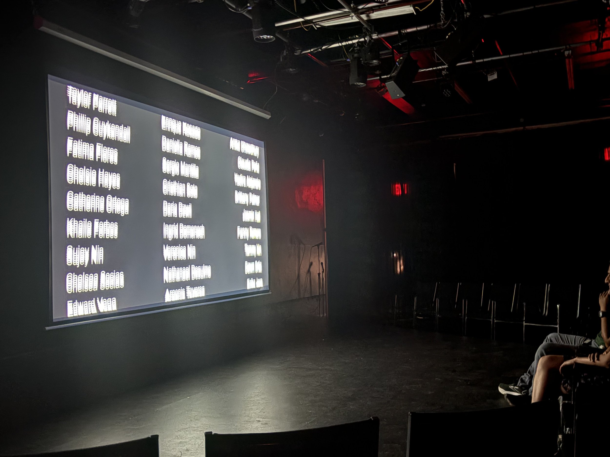 A dark indoor space with a large illuminated screen displaying names of people, with some chairs and two people sitting on the right.