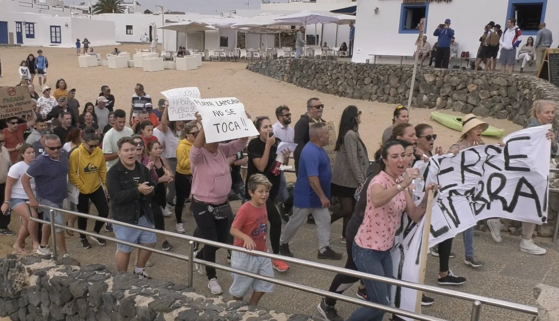 Group of protesters marching along a pathway on a beach, holding signs and banners, with a white building and rocky wall in the background.