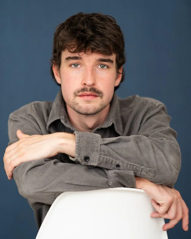Portrait of a young man with brown hair and a mustache, wearing a gray button-up shirt, leaning on a white chair against a solid blue background.