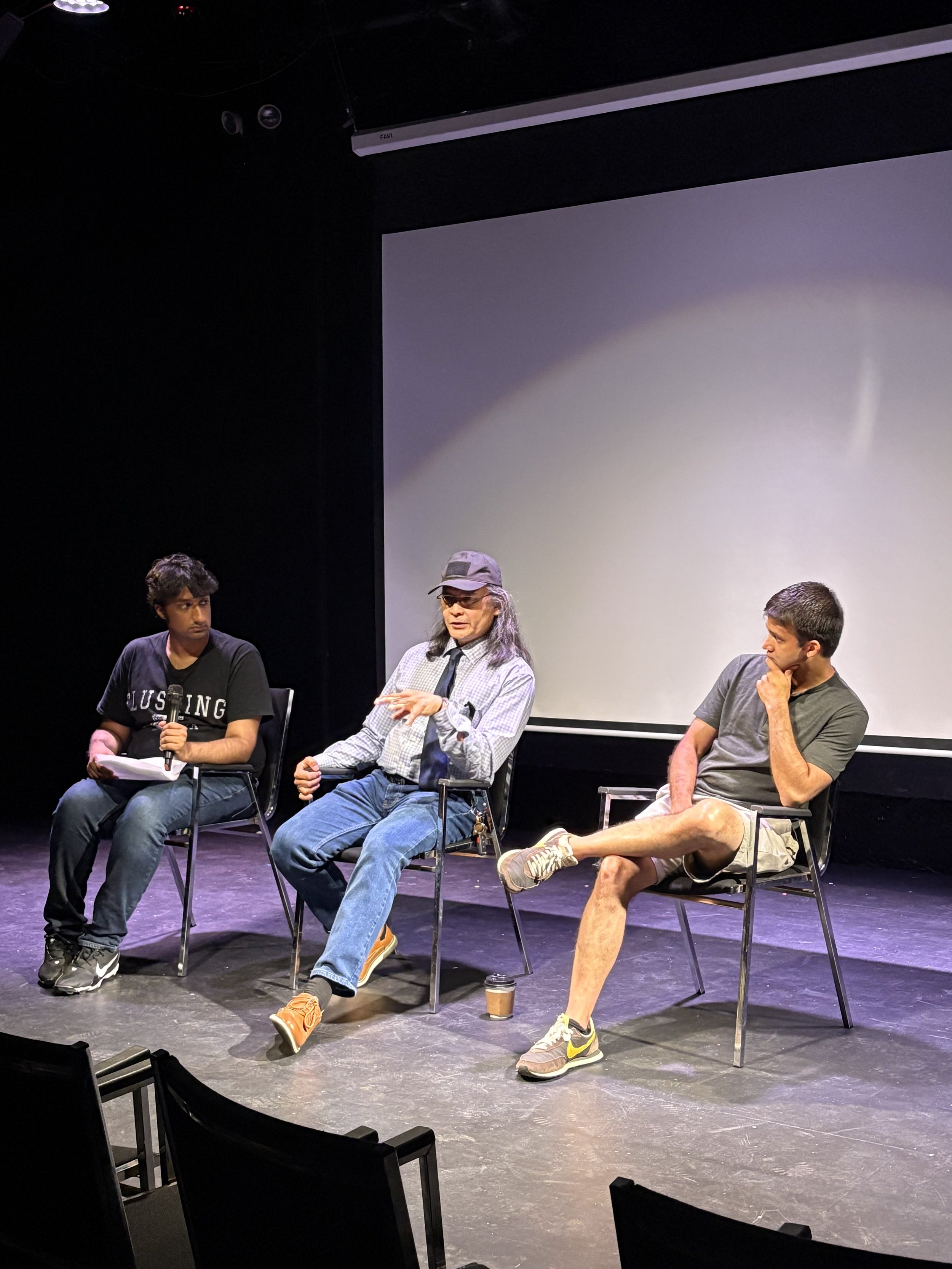 Three men seated on a stage, engaging in a discussion or panel. The man in the middle is wearing glasses, a gray cap, and casual clothing, gesturing with his hand. The man on the left is holding a microphone and notes, dressed in a black t-shirt and jeans. The man on the right is wearing a gray t-shirt and shorts, sitting with his legs crossed and one hand on his chin. A large blank screen is behind them, and there are empty chairs in the foreground.