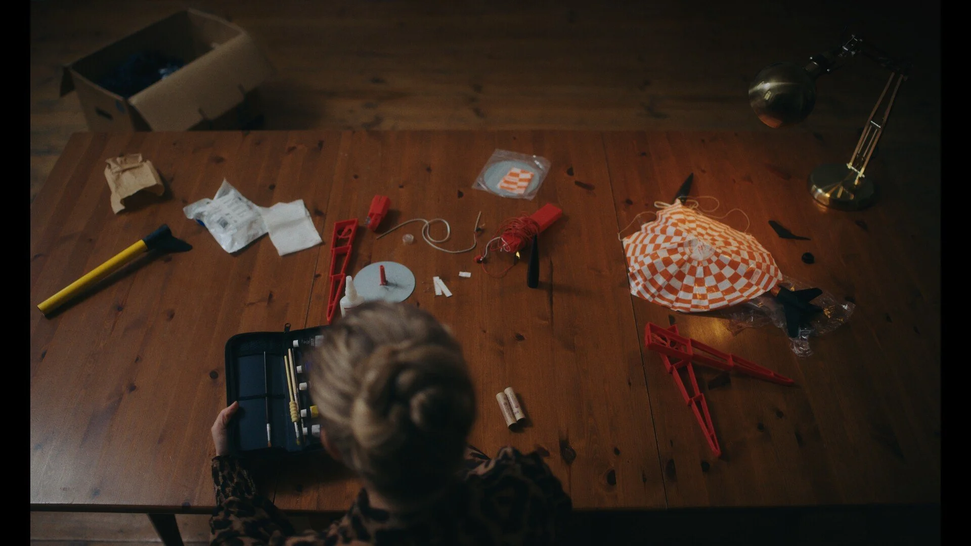 Child with blonde hair working with orange and white checkered paper lantern craft on a wooden table, surrounded by craft supplies and tools.