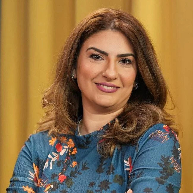A smiling woman with shoulder-length brown hair, wearing a blue floral blouse, standing in front of a yellow curtain.