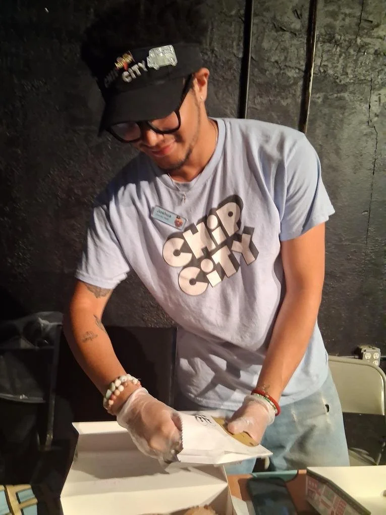 A young man wearing a black cap, glasses, and a gray t-shirt that says 'Chirp City' is opening a box of food with gloves. He is standing in front of a black wall in what appears to be a casual setting.