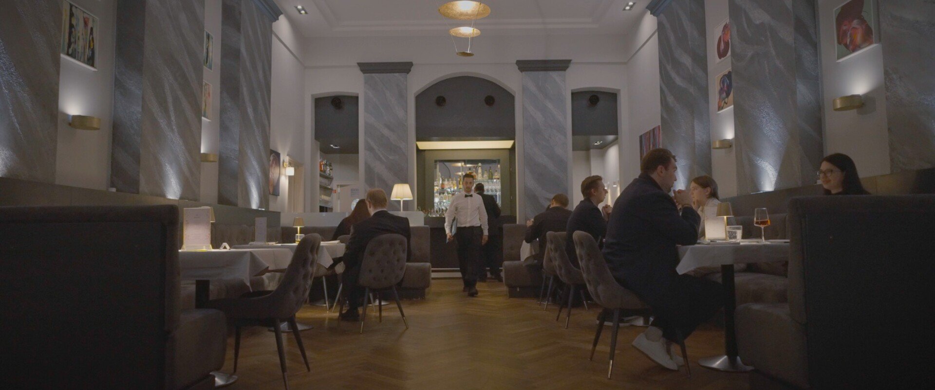 Elegant restaurant interior with diners seated at tables, waitstaff serving guests, and sophisticated decor featuring paintings and ambient lighting.