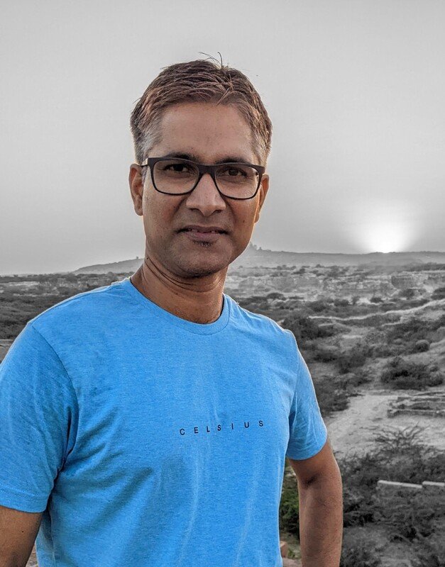 A man with short brown hair and glasses wearing a blue T-shirt, standing outdoors with a landscape of hills and a cloudy sky in the background.