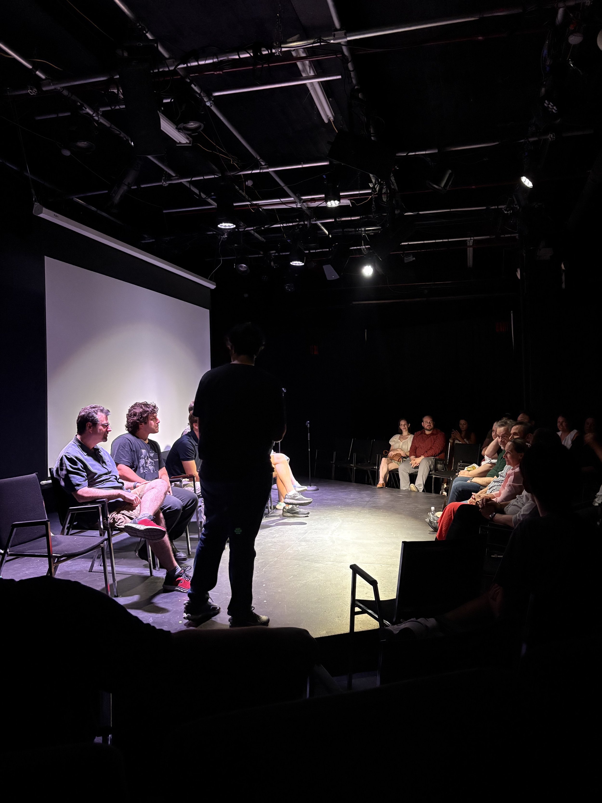 People participating in a panel discussion on a small stage in a dimly lit theater, with an audience seated around the stage.