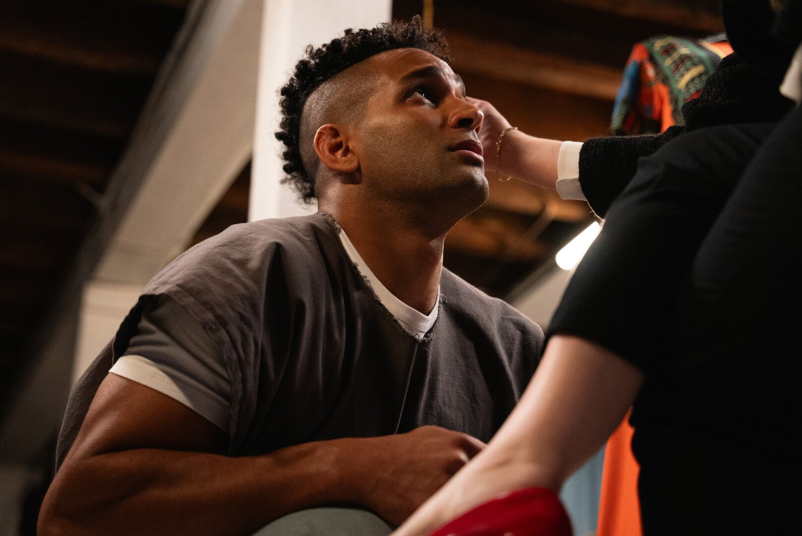 A young man with curly hair and a shaved side hairstyle looks up while a woman touches his face. The scene appears to be in a room with wooden beams.