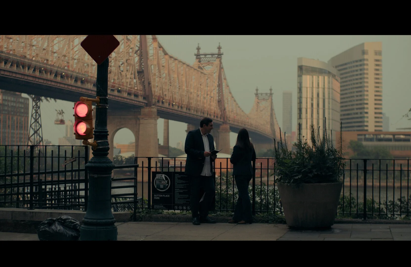 Two people talking on a sidewalk near a fence with a large planter, with a bridge and tall buildings in the background, under a red traffic light.