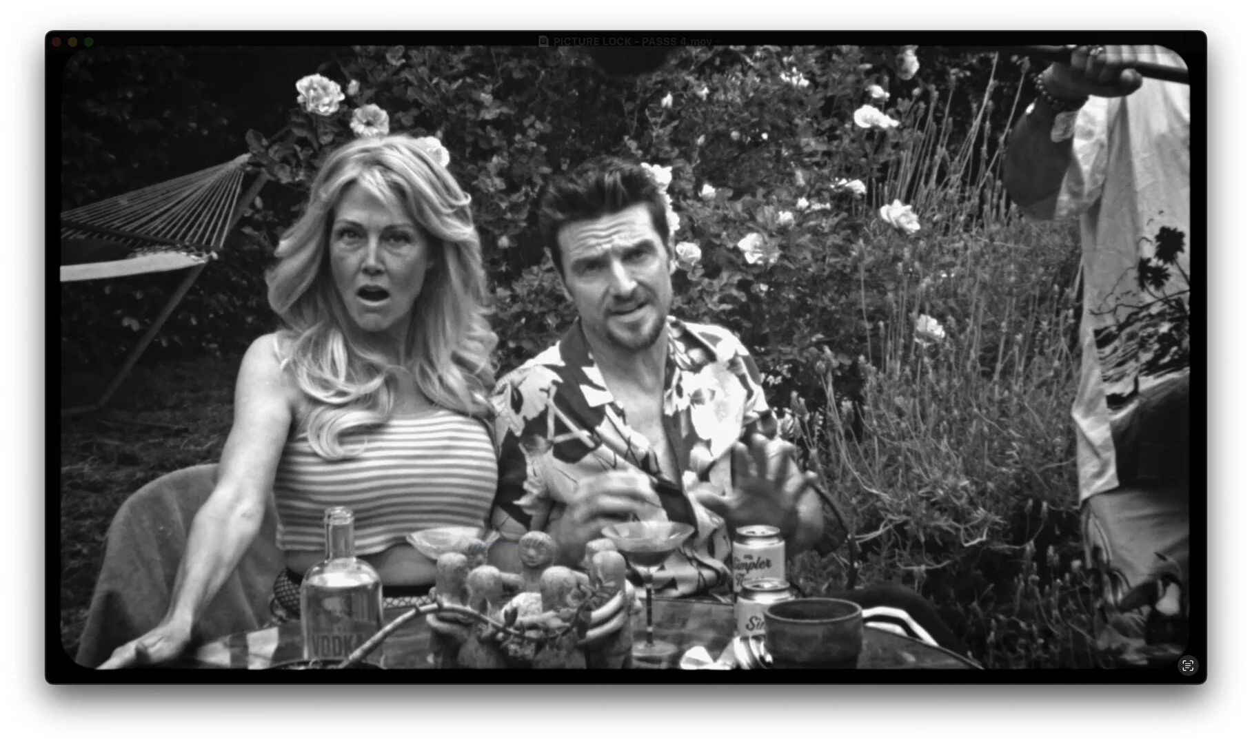 A black and white photo of a man and woman sitting at an outdoor table with food and drinks, with a floral garden background and a person standing on the right.