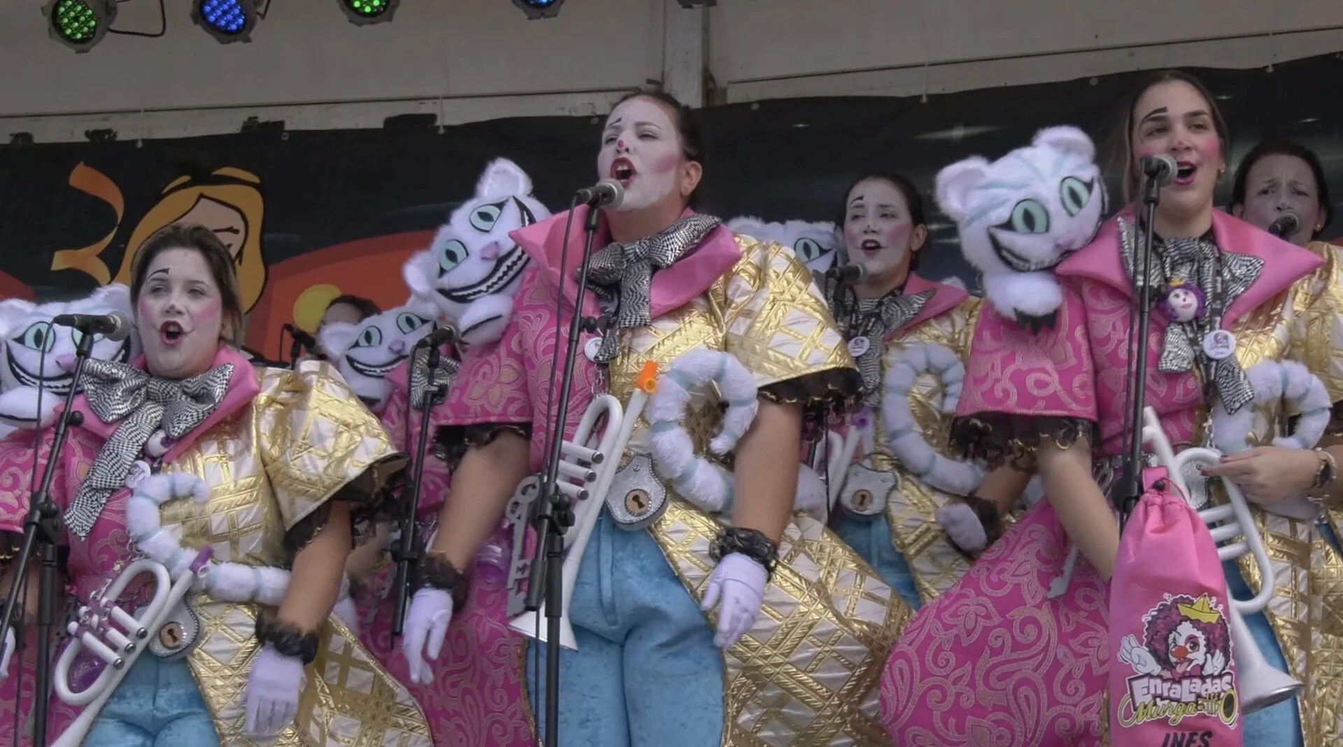 Group of women dressed as characters from the musical 'Cats' performing on stage, wearing colorful costumes with pink, gold, and blue patterns, and waving white plush props shaped like cats