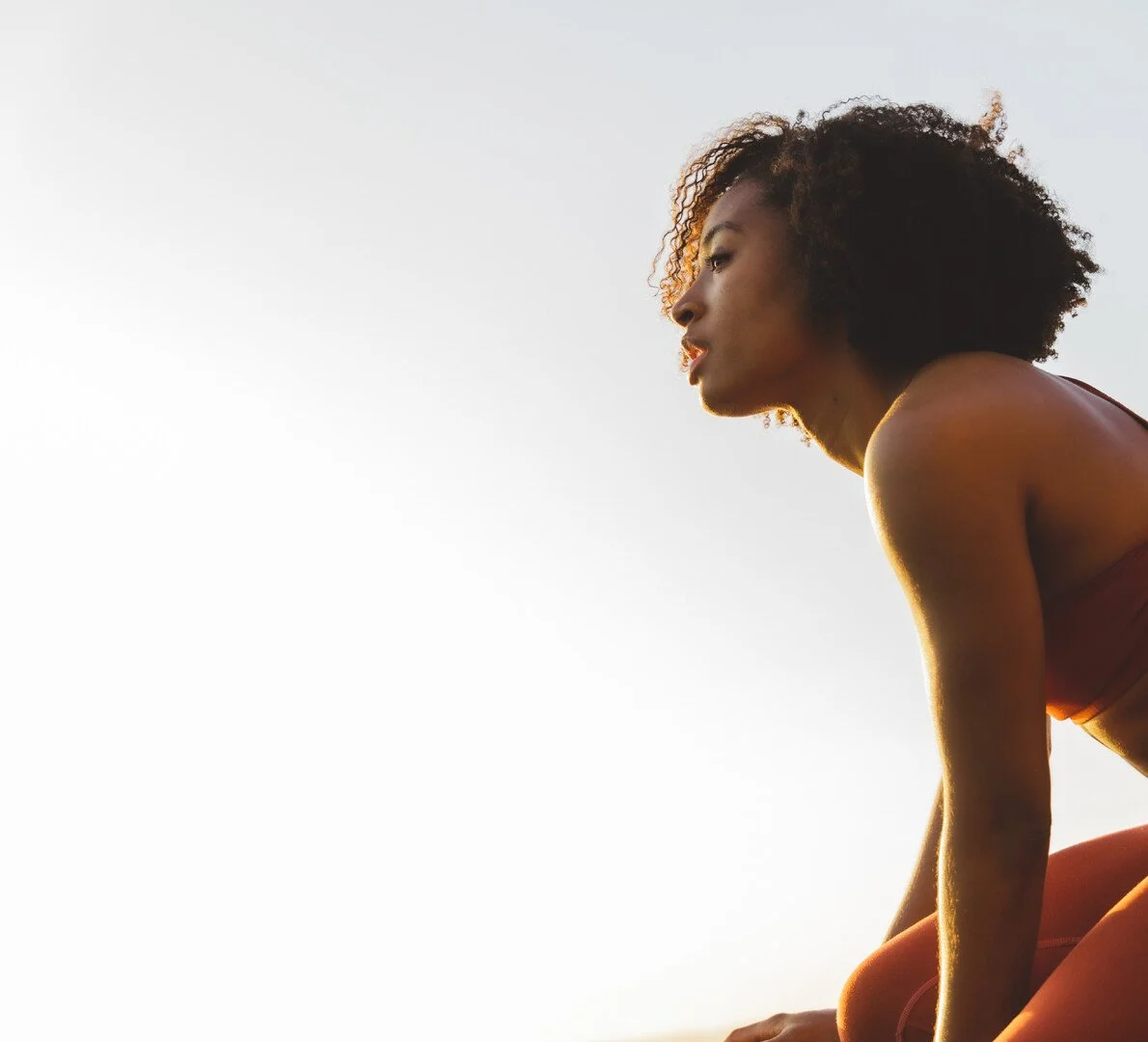 A woman with curly hair and wearing activewear is in a stretching or yoga pose outdoors during sunset with a clear sky.