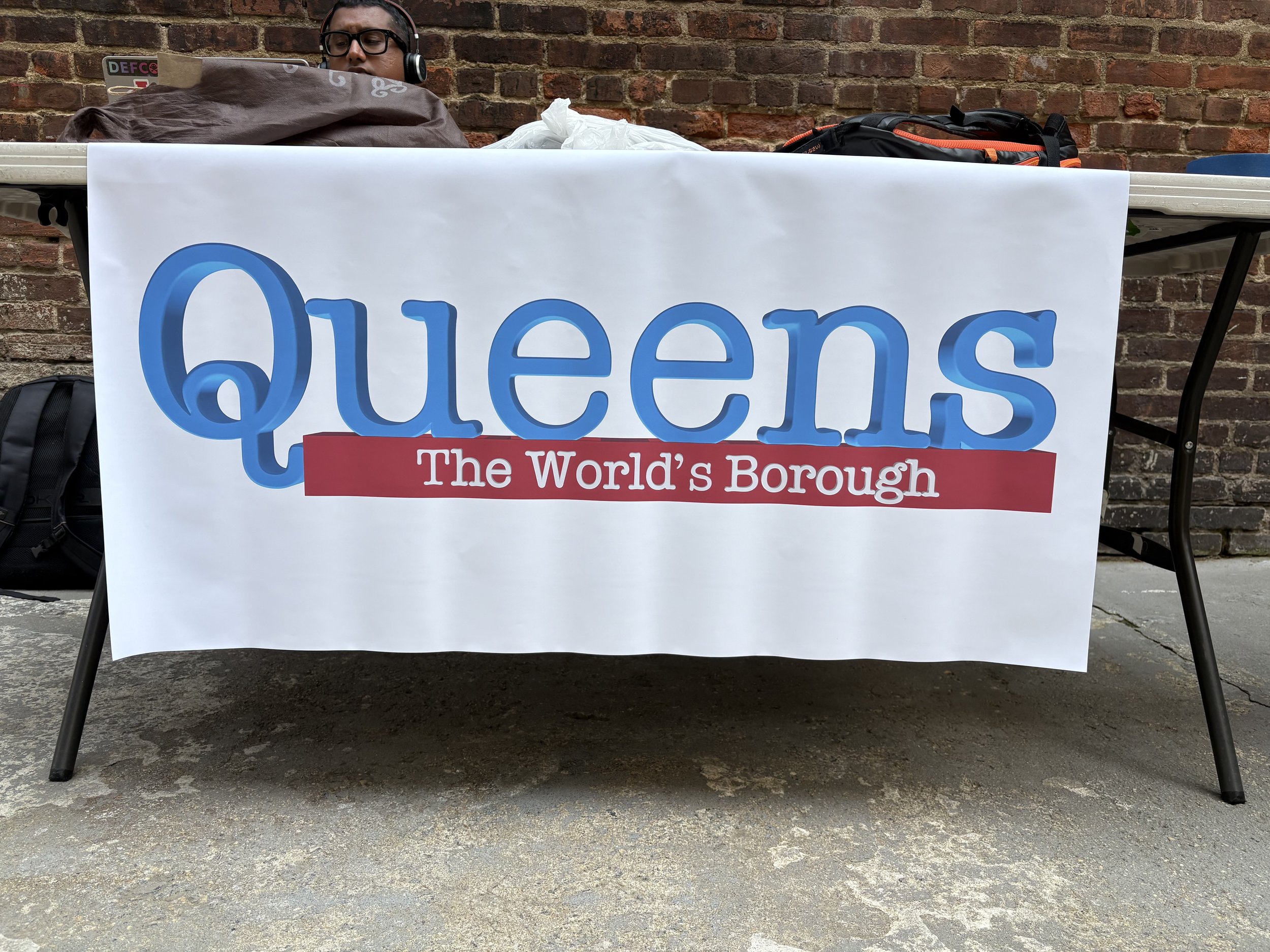 A white banner on a table reading "Queens" in large blue letters with a tagline "The World's Borough" in white letters on a red background, set against a brick wall. There is a person partially visible behind the banner, wearing glasses and headphones, working at a laptop.