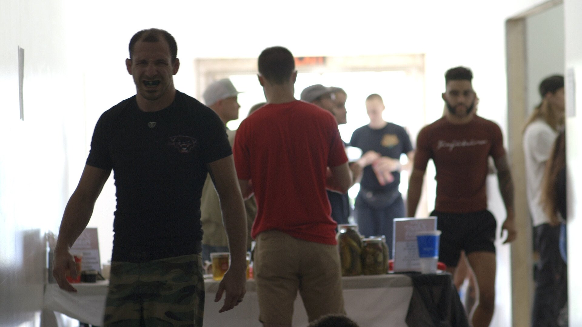 Group of people socializing indoors, some sitting at a table with jars and drinks, bright light from open door in background, casual attire.