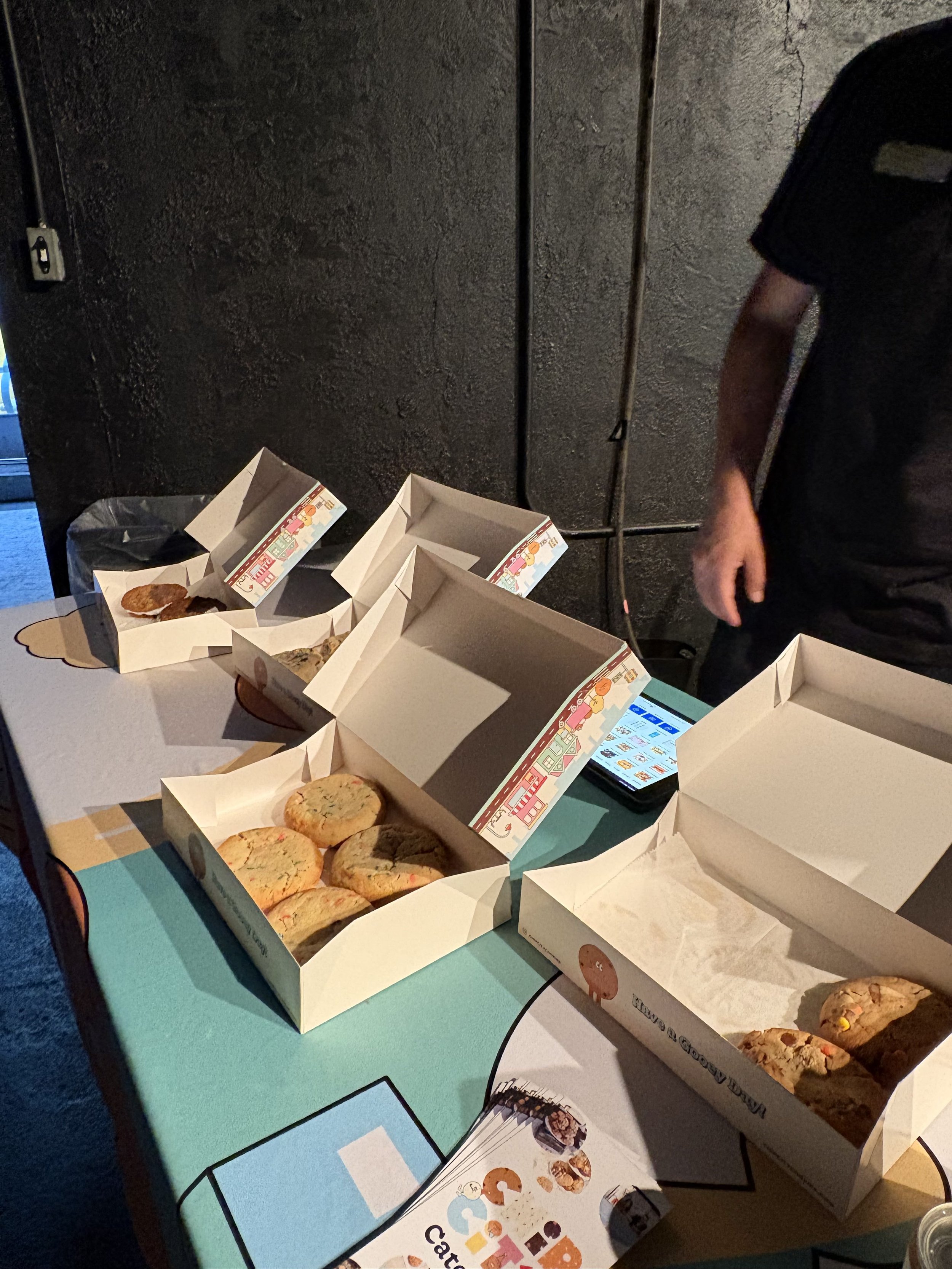 Several opened bakery boxes with cookies and pastries on a table, with a person standing nearby and a smartphone visible.
