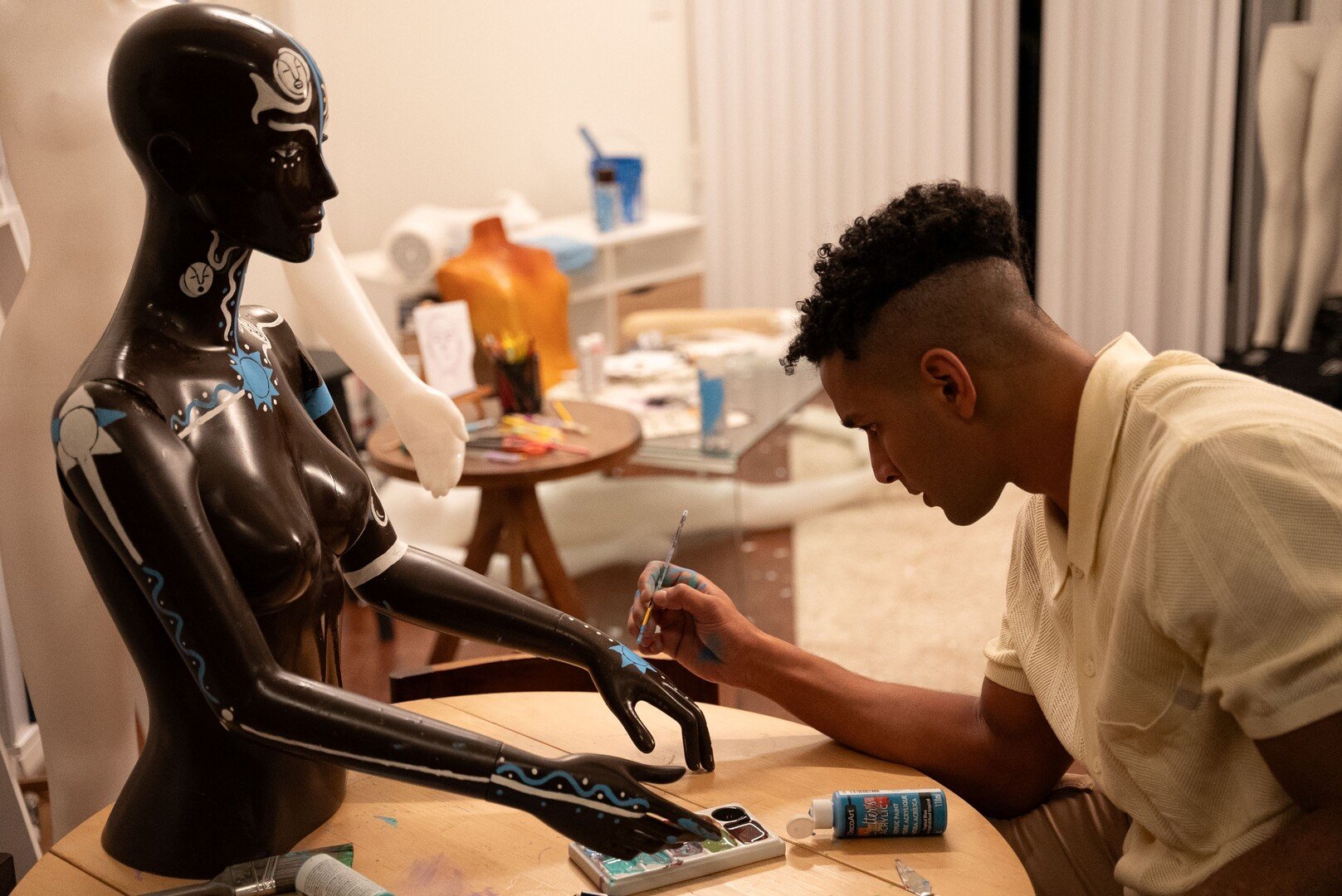 An artist paints blue and white designs on a black robot sculpture of a woman, working at a wooden table in a well-lit room.