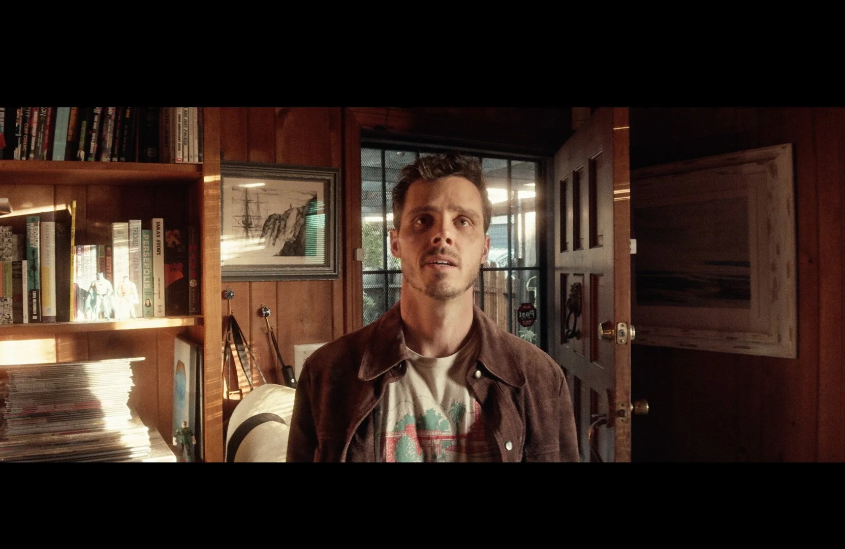 A man with brown hair and a mustache wearing a graphic t-shirt and brown jacket standing inside a wooden-paneled room with a bookshelf, framed picture, and open door behind him.