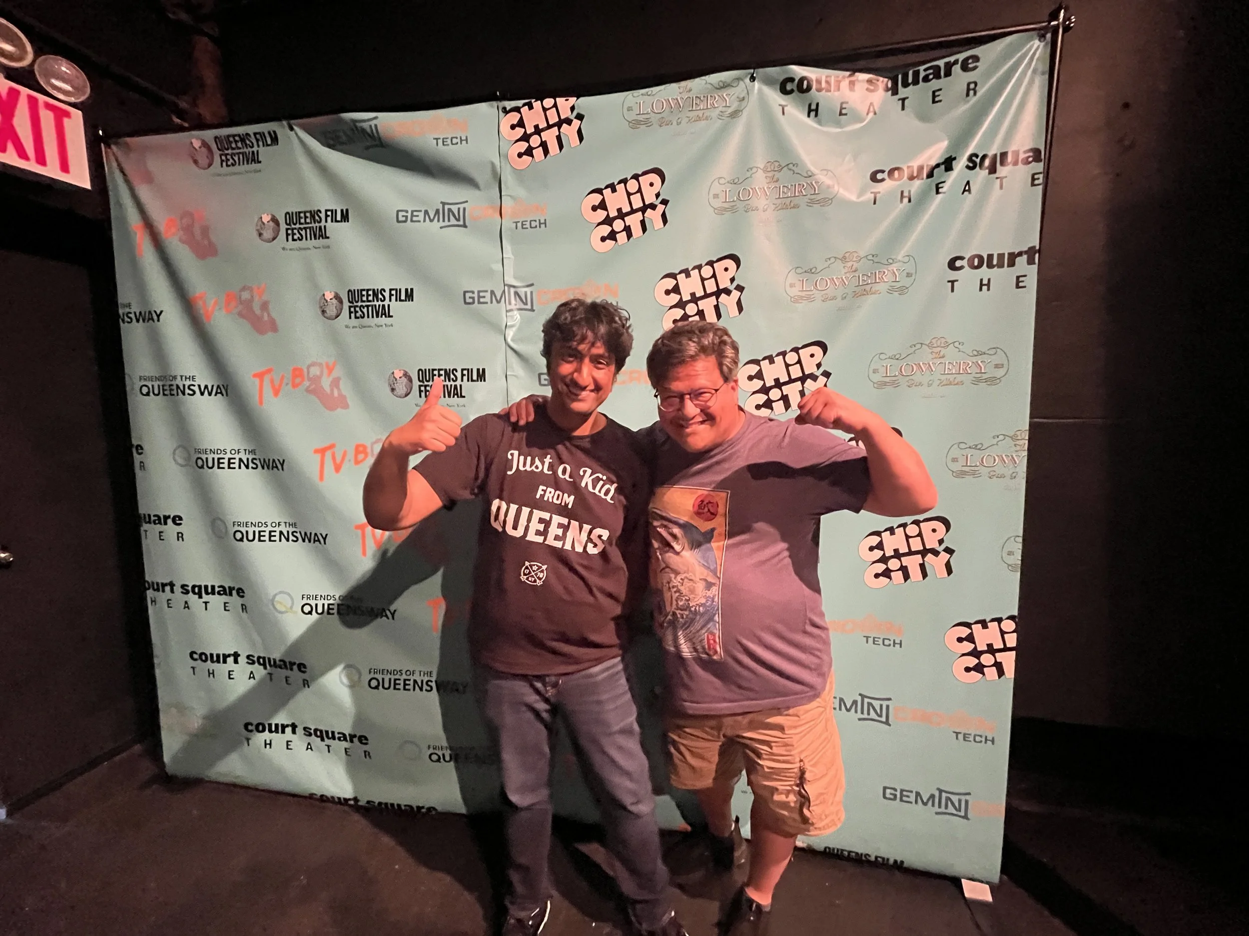 Two men standing next to each other and flexing their arms in front of a backdrop with event logos. One of the men is wearing a black T-shirt with white text that reads "Just a Kid from QUEENS," and the other man is wearing a gray T-shirt with a graphic design. Both are smiling, and the backdrop features logos for Chip City, Queens Film Festival, Queen's Way, Court Square Theater, Lowery, and GEMINI.