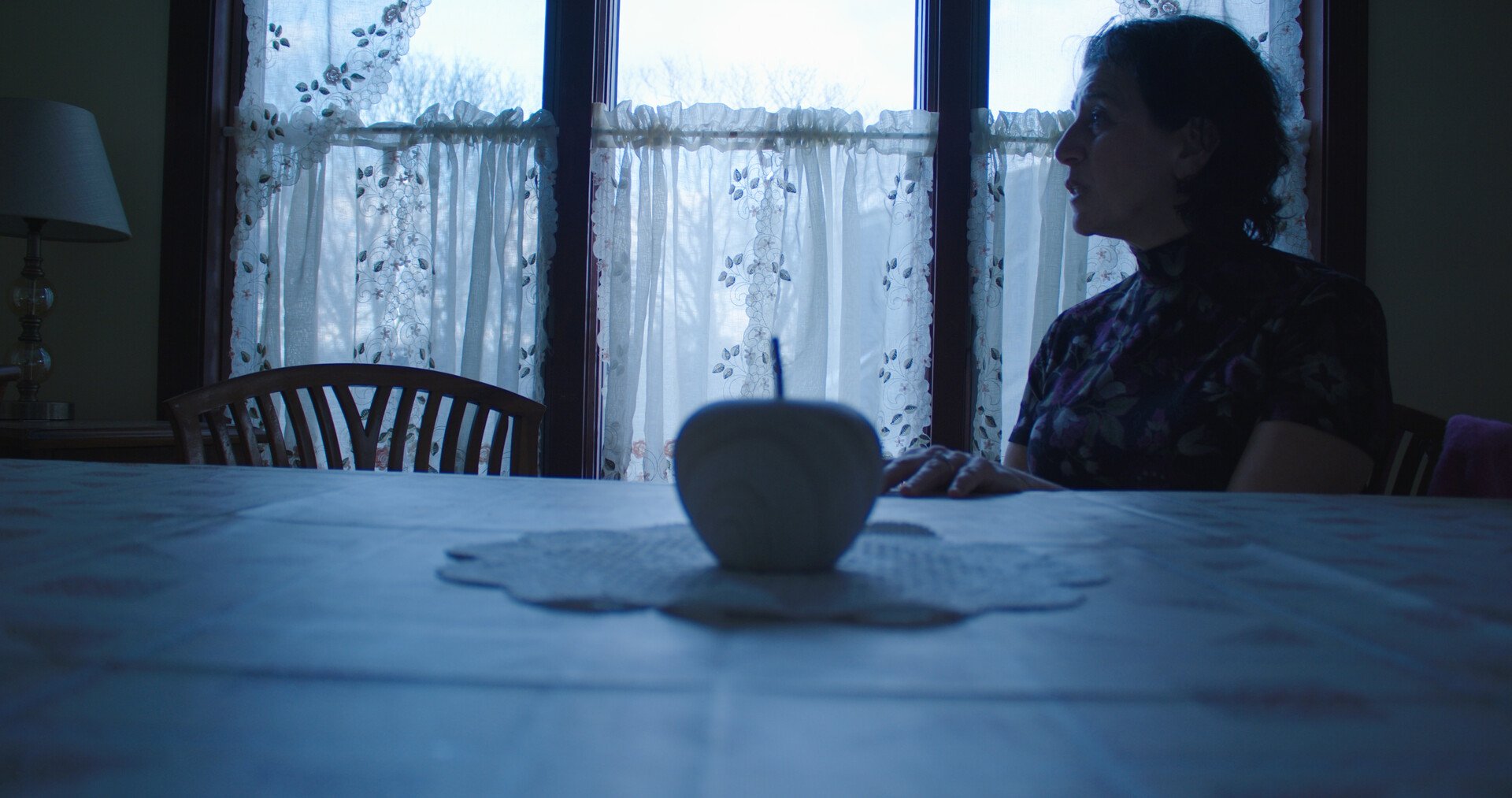 A woman sitting at a table by a window with lace curtains, with an apple on the table in the foreground